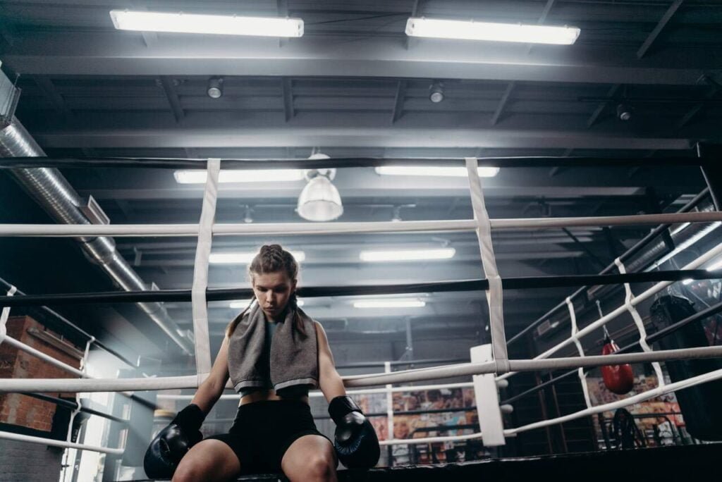 Odkrywanie własnej ścieżki życia, kobieta w rękawicach bokserskich i z ręcznikiem przewieszonym na karku siedzi smutna na skraju ringu
